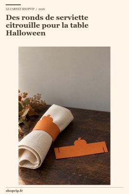 Serviette écrue roulée dans un anneau de carton orange à silhouette de citrouille ; une bande ouverte posée à plat montre les fentes de montage.